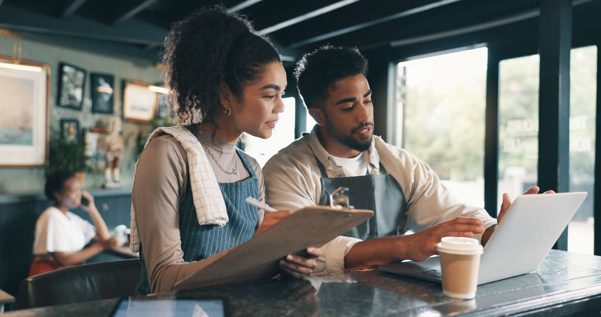 Two coworkers collaborate at a cafe counter: a woman with a clipboard and a man with a laptop and a coffee cup nearby.