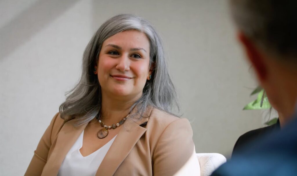 Smiling woman with silver hair wearing a beige blazer, engaged in a conversation with someone off-camera.