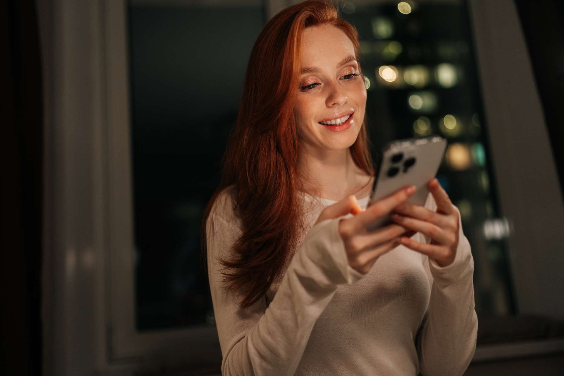 Smiling red-haired woman looking at her smartphone at night, indoors, in a cozy setting.