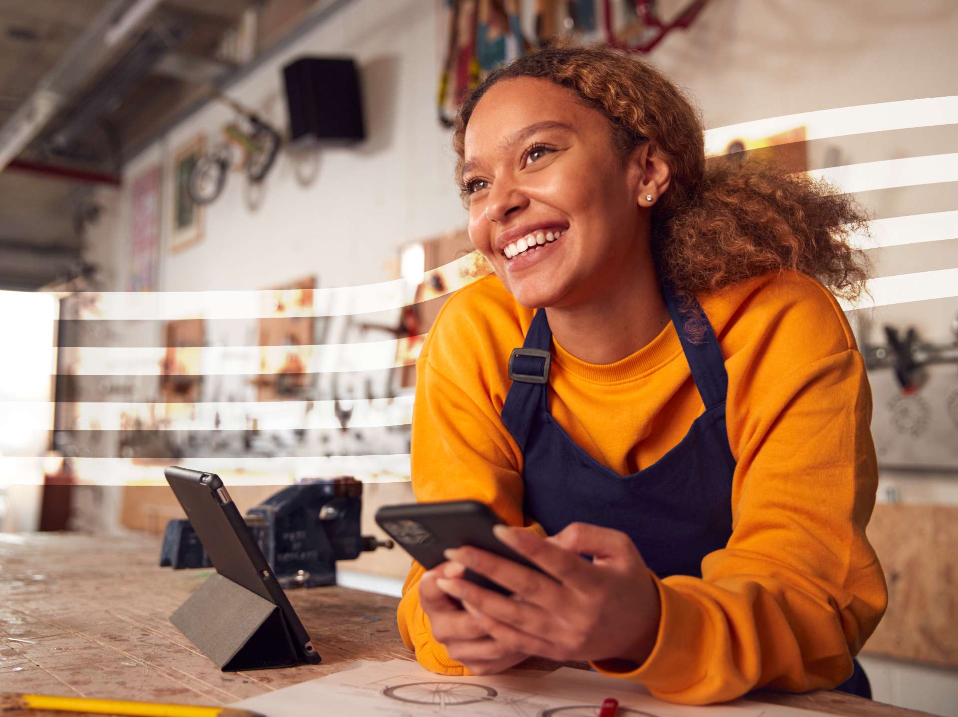 Worker in an orange shirt and blue apron, smiling while using a smartphone in a busy workshop.