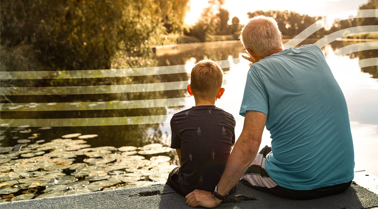 Grandfather and grandson sit together on a lakeside ledge at sunset, looking over a pond with lily pads.